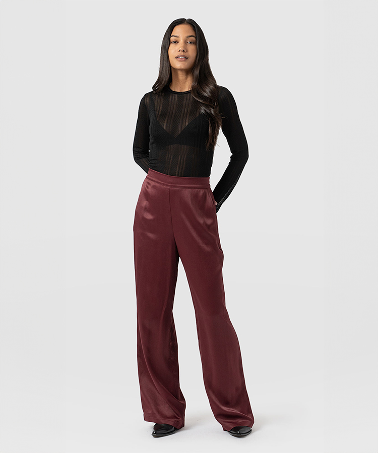 A model wearing a sheer black long sleeve top and maroon satin trousers