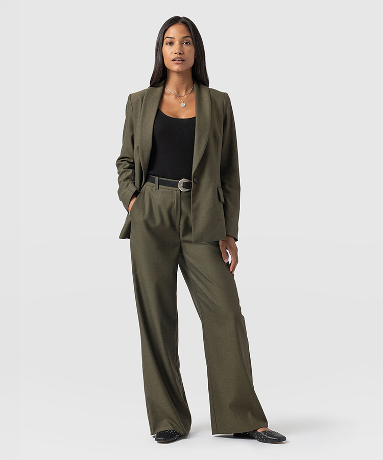 A model wearing an earthy green suit with a single-breasted blazer and wide leg trousers. Layered over a black tank top with a black leather belt.