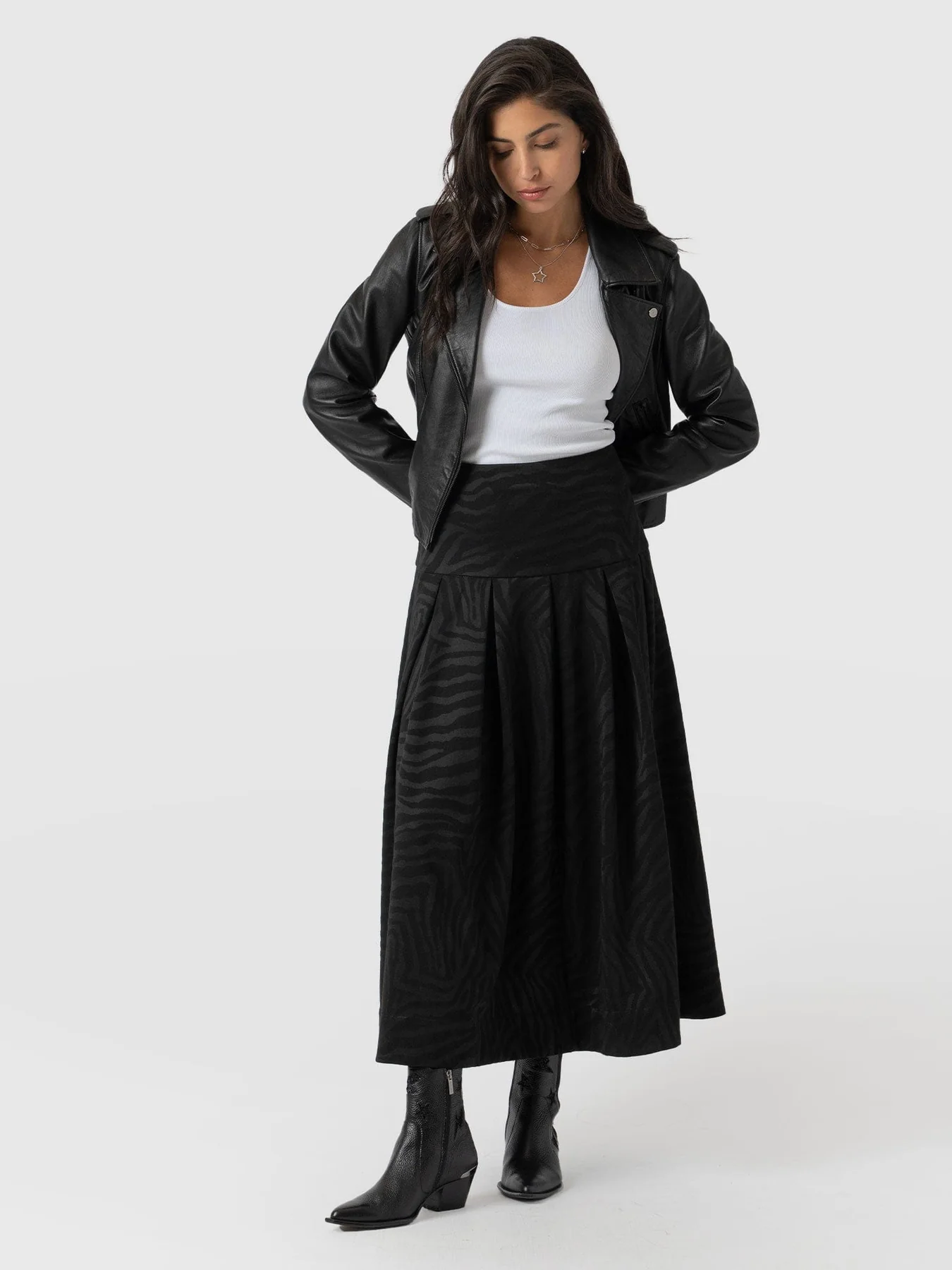 A model wearing a leather jacket, a white tank top, a black-on-black zebra print midi skirt, and the Billie Rock ankle boots.