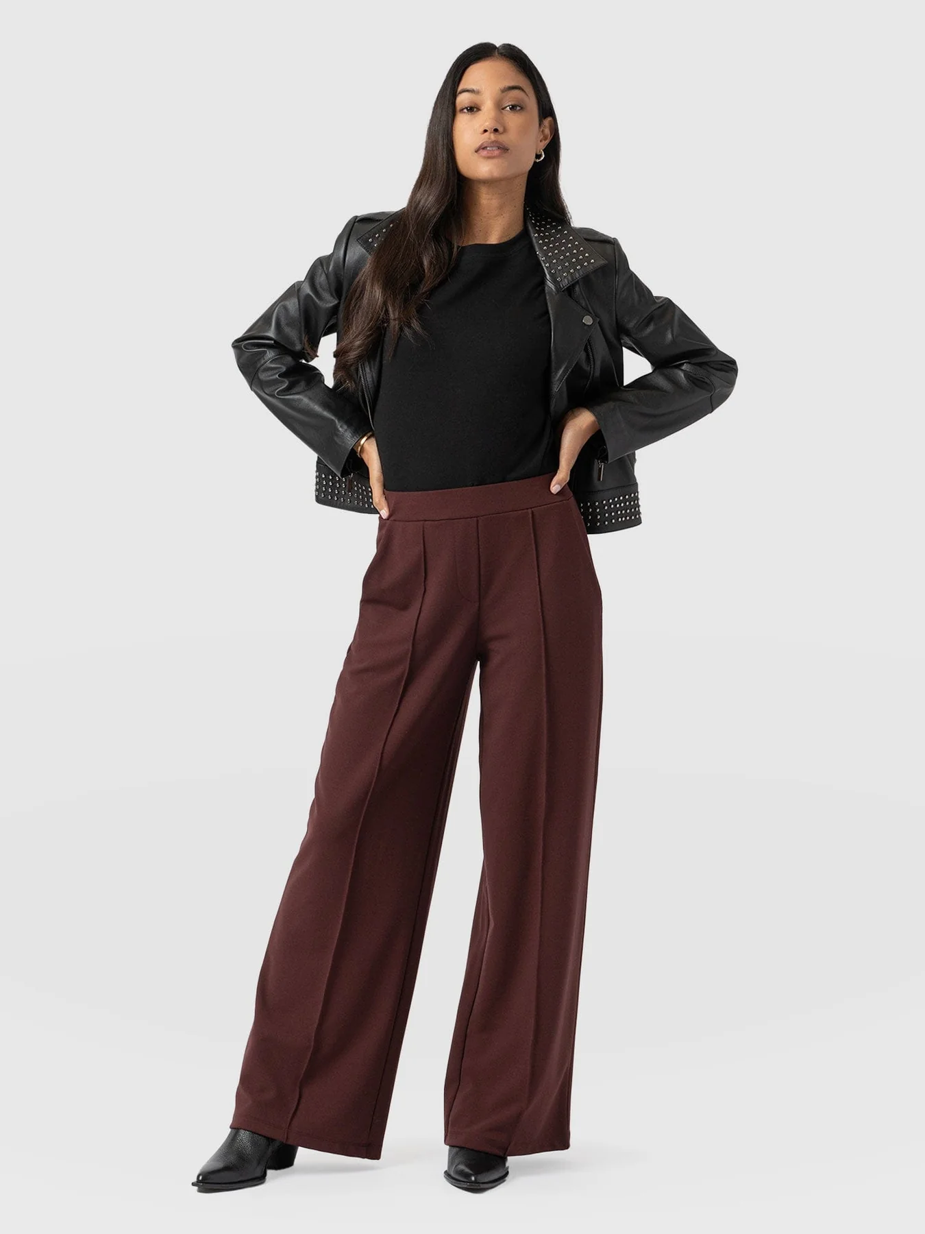 A model wearing a leather jacket, a black T-shirt tucked into burgundy wide leg trousers, and the Saint and Sofia Billie Rock ankle boots.