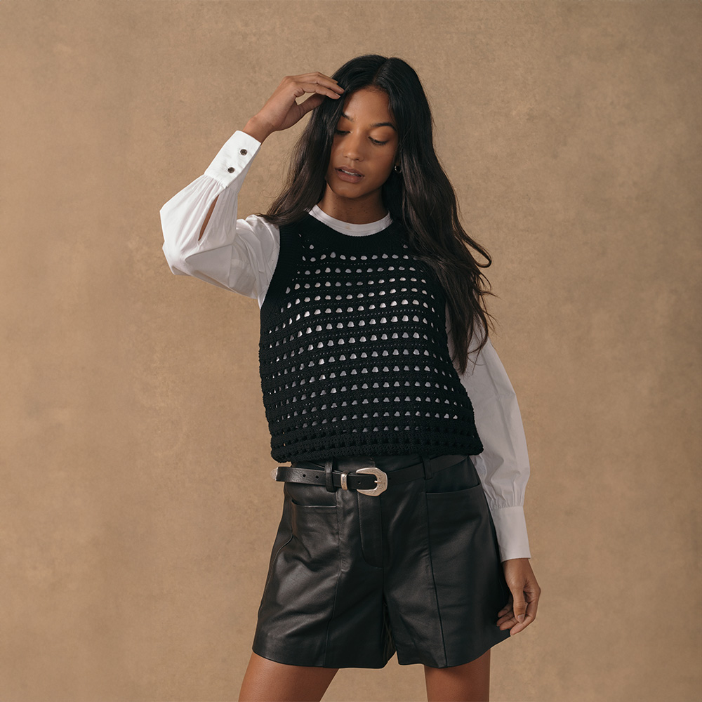 A model wearing a black sleeveless knitted vest over a white long-sleeved shirt and black leather shorts.