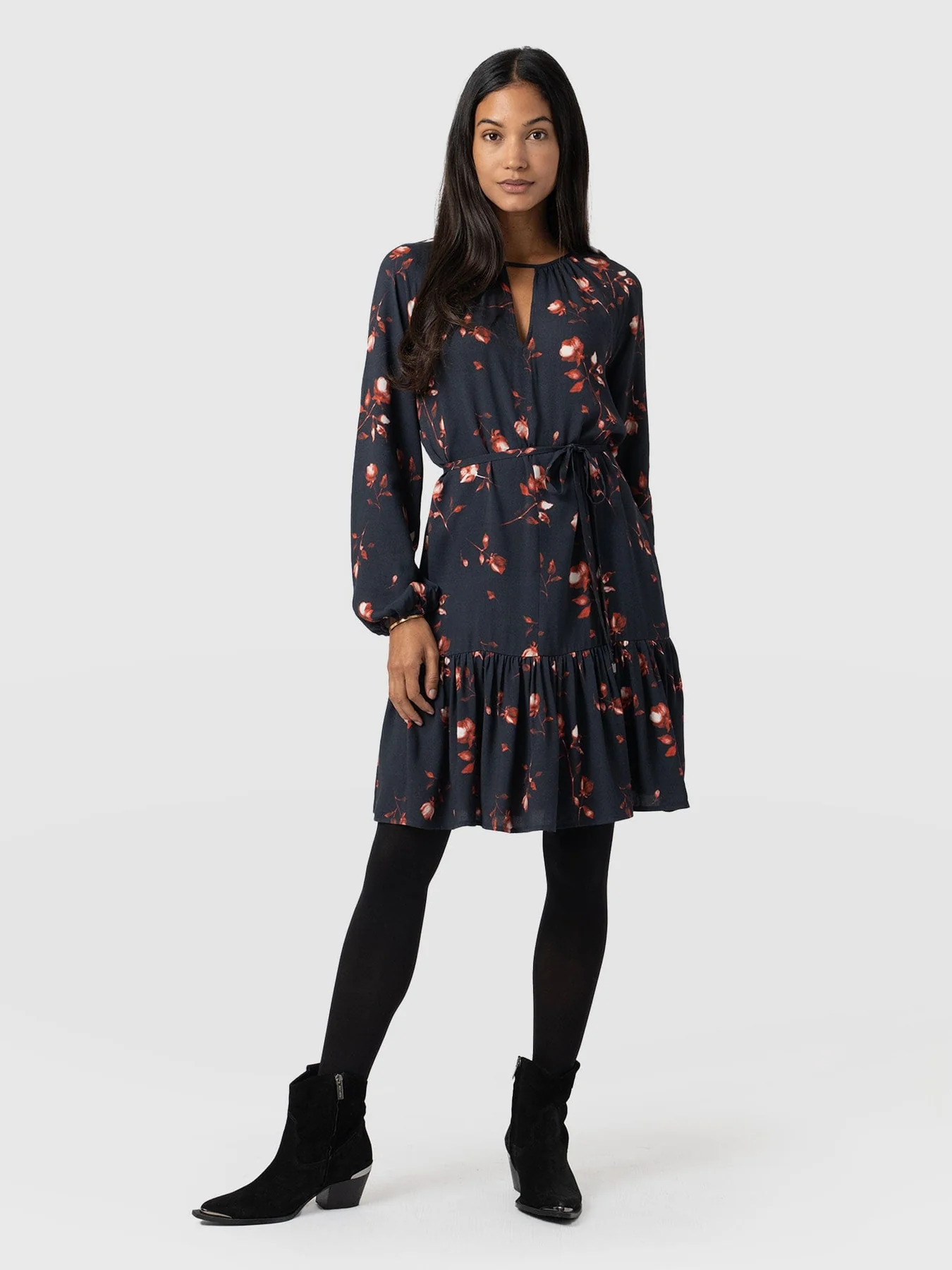 A model wearing the Fia Mini Dress from Saint and Sofia's November collection. The floral dress is styled with black tights and black suede ankle boots.
