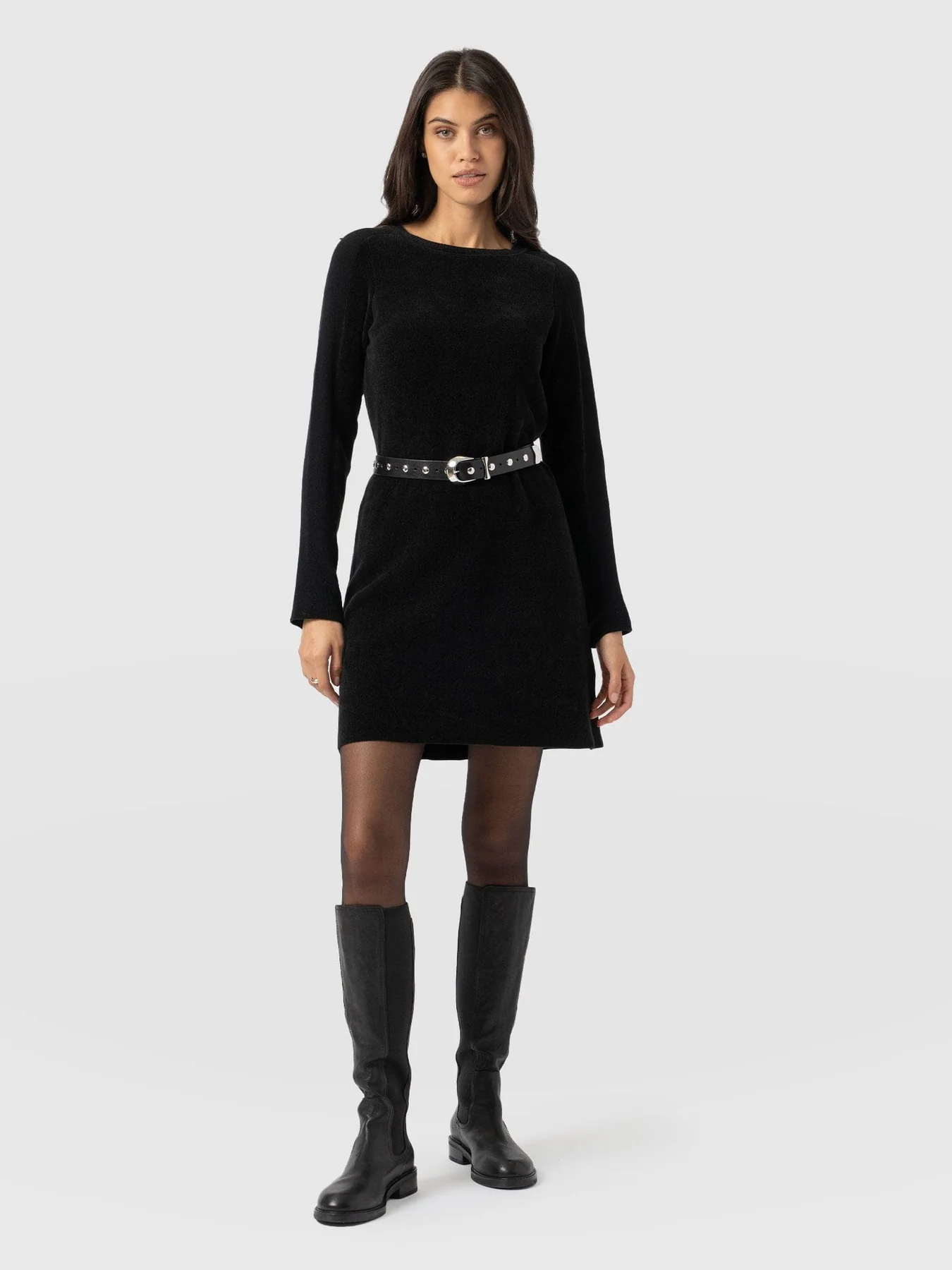 A model wearing the Raya Knit Dress from the Saint and Sofia November collection with a studded black belt around the waist, sheer black tights, and knee-high leather boots.