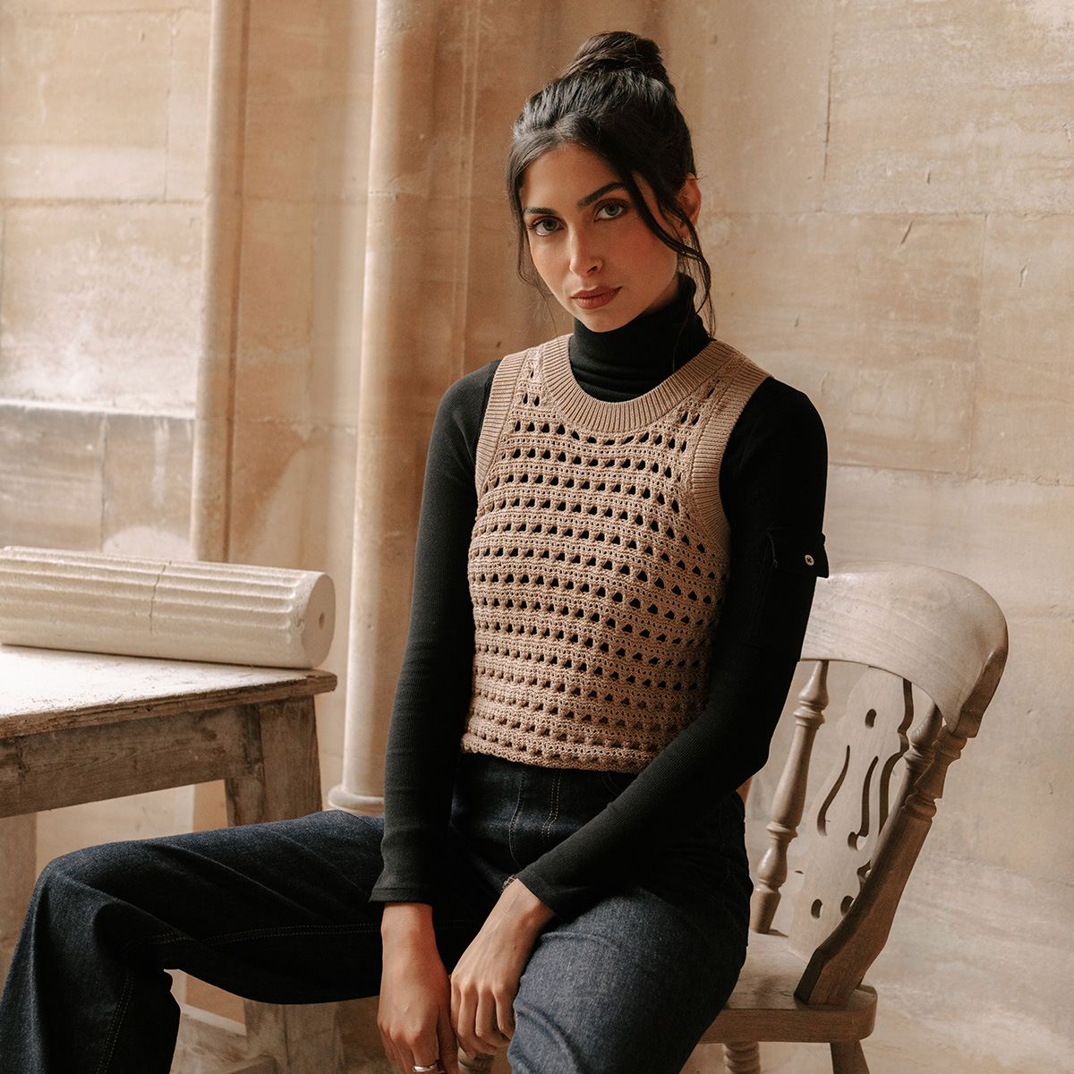 A model sitting on a chair wearing a black turtleneck with a brown knitted vest layered over the top.