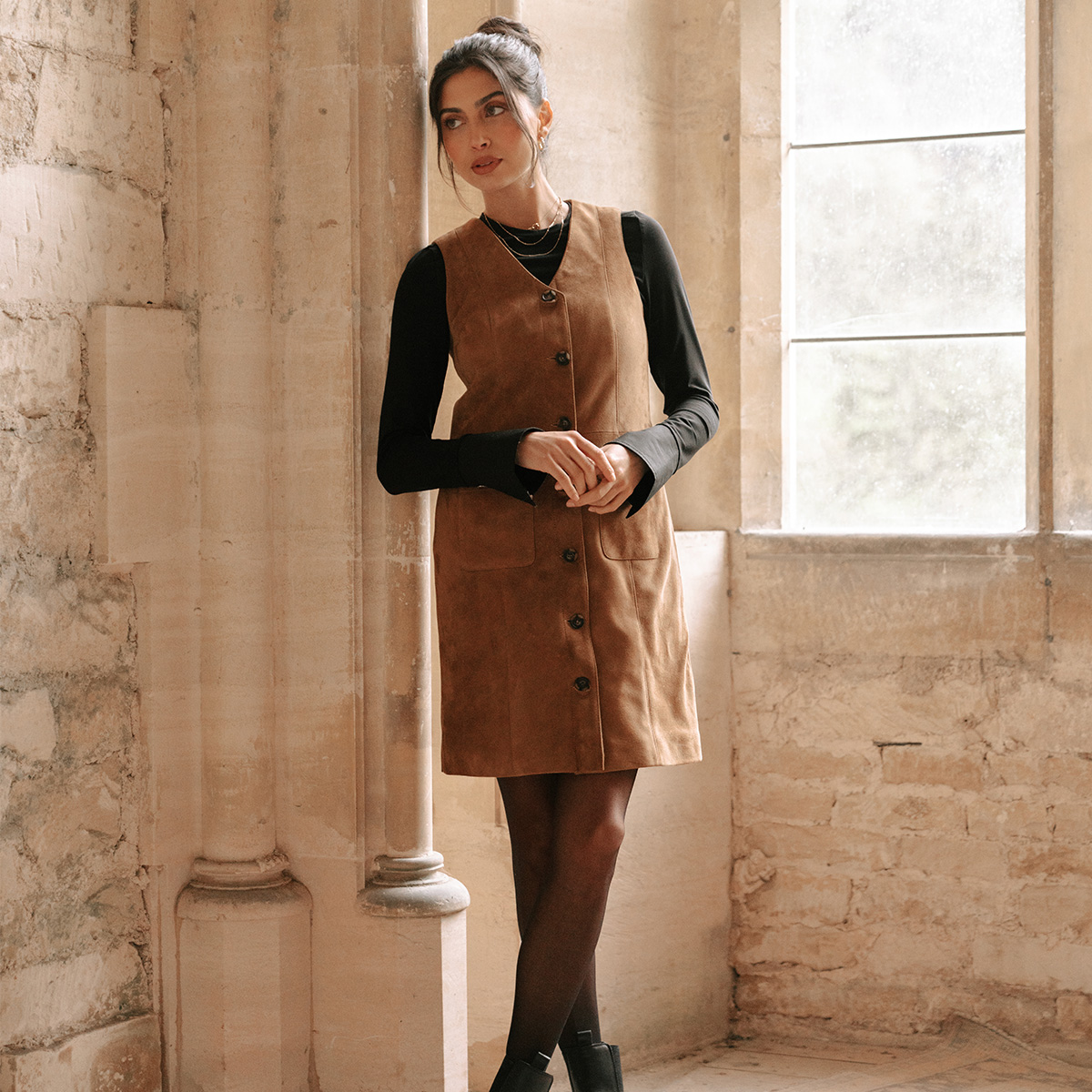 A model wearing a black long sleeve tee layered under a suede pinafore dress with sheer black tights and black ankle boots.