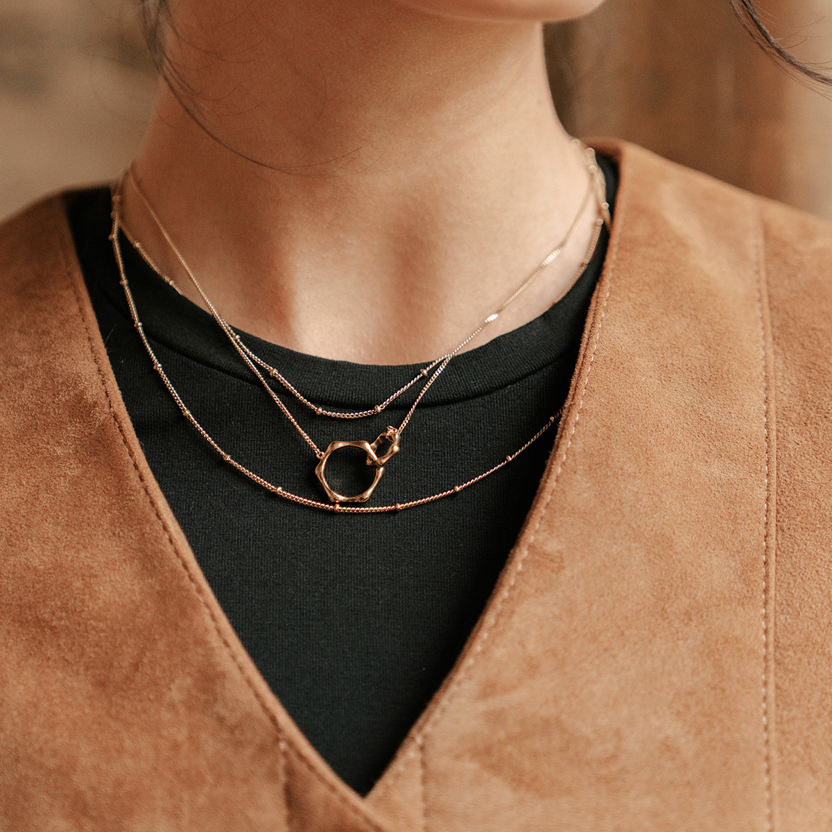 A model wearing a black long tee layered under a tan suede pinafore dress with layered gold necklaces.