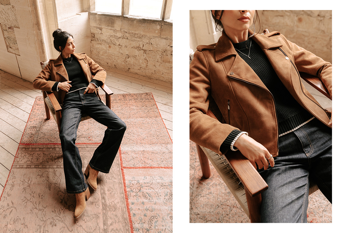 A model wearing a Saint and Sofia brown suede jacket with a black knitted jumper and dark blue denim flared jeans. She's sat on a wooden chair that sits on a red rug.