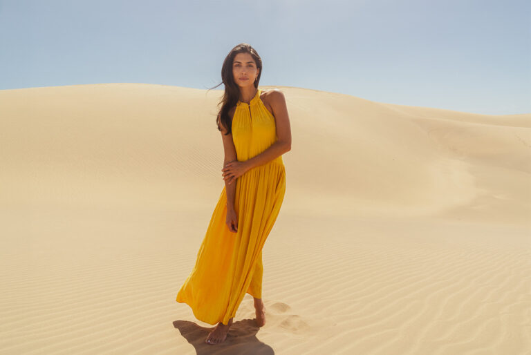 A model stood in the centre of a sand dune backdrop wearing a yellow halterneck maxi dress