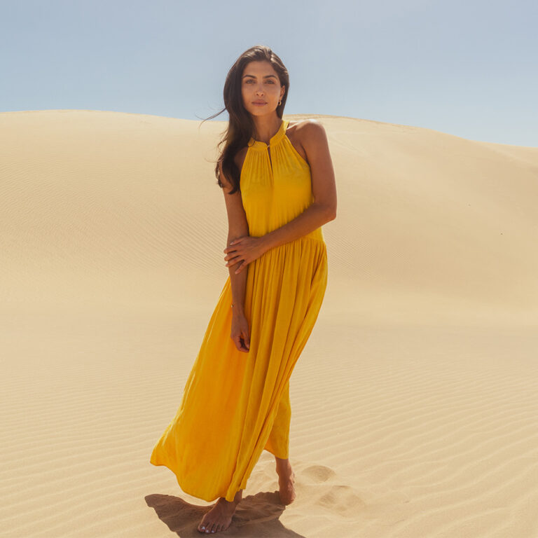 A model stood in the centre of a sand dune backdrop wearing a yellow halterneck maxi dress