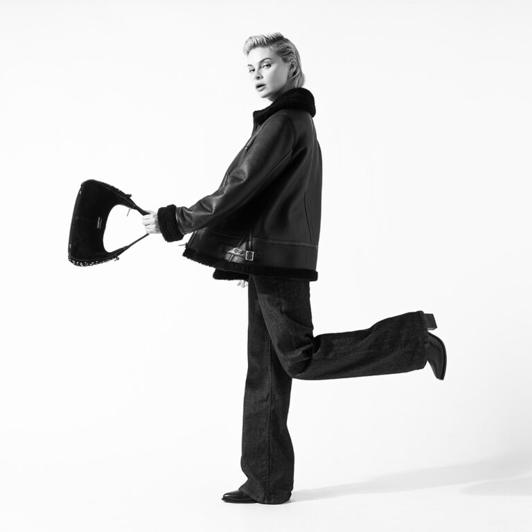 A black and white image of a model wearing a Saint and Sofia aviator jacket, a pair of black jeans, and heeled boots. She is stood to the side with one leg kicked out behind her and swinging a handbag in front of her.