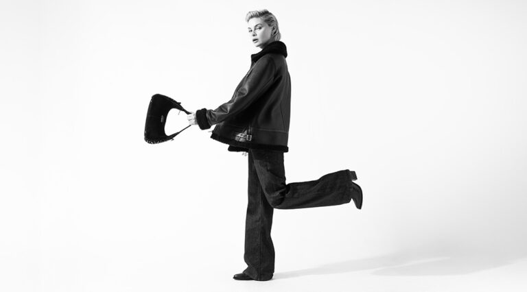A black and white image of a model wearing a Saint and Sofia aviator jacket, a pair of black jeans, and heeled boots. She is stood to the side with one leg kicked out behind her and swinging a handbag in front of her.