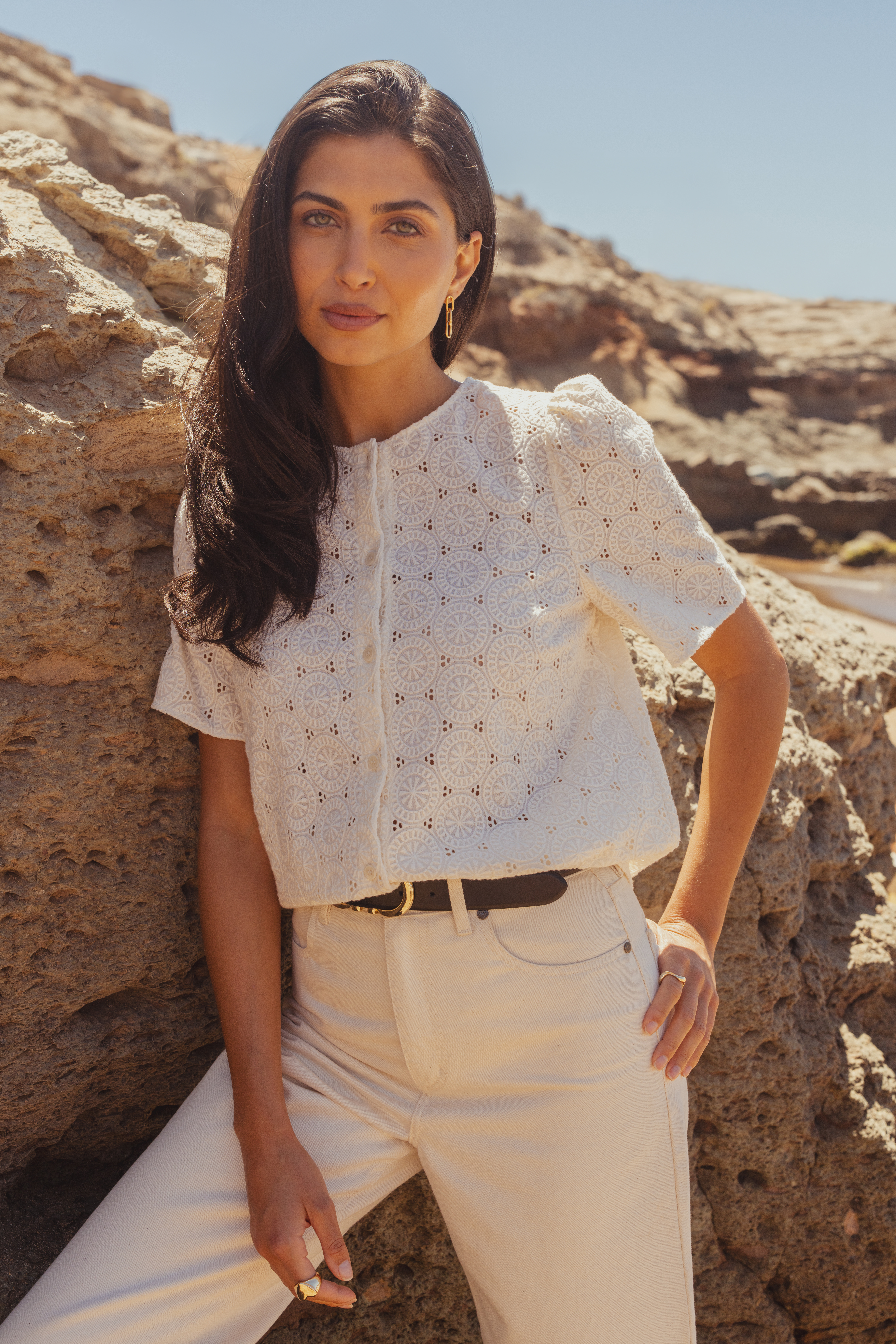 A model wearing the Saint and Sofia Tabitha Blouse. A short-sleeved broderie anglaise design with puffed sleeves and a button up fastening. It's styled with cream barrel leg jeans with a leather belt.