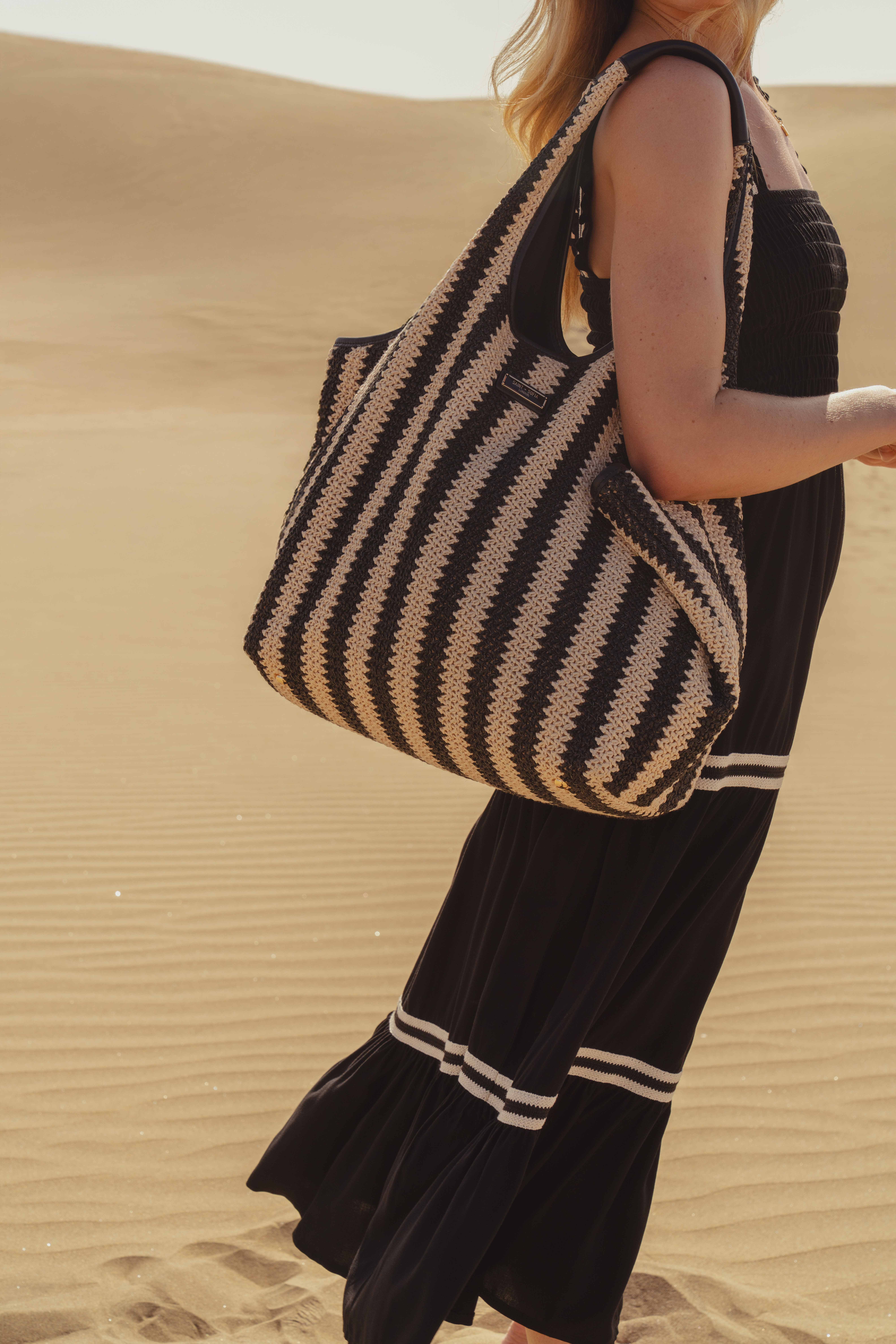A model wearing a black dress with the Saint and Sofia Stella Shoulder Tote on her shoulder. This boho style bag is a black and beige raffia style with two top handles and a roomy interior.