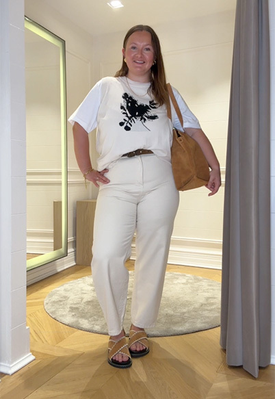 A size 14 model wearing the Saint and Sofia Astra sweater vest layered over a short sleeve crew neck tee and paired with white barrel leg jeans and sliders for a spring look.