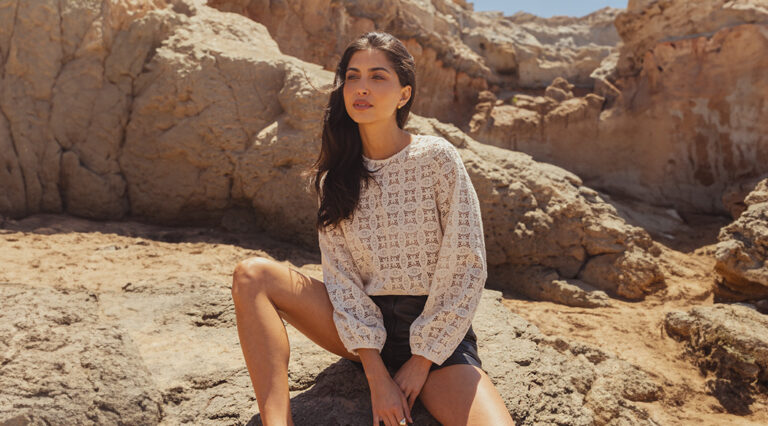 A model sat on a rocky terrain wearing a Saint and Sofia broderie anglaise long sleeve blouse with leather shorts.