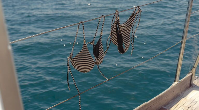 A striped bikini by saint and sofia hanging from the railings of a boat with the ocean as the backdrop