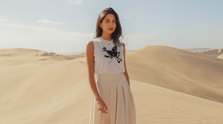 A model standing in the dunes wearing the Saint and Sofia Astra Sweater Vest with a linen midi skirt