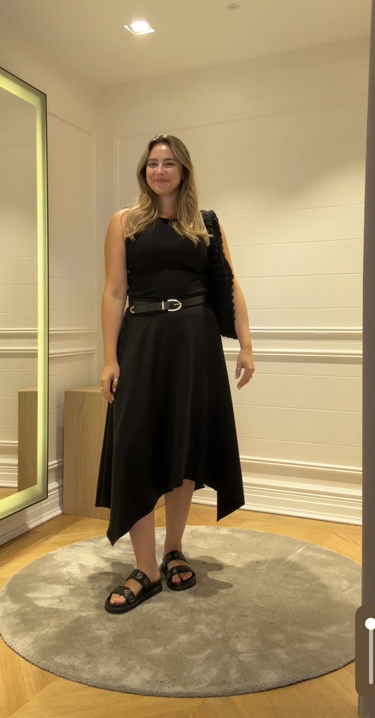 A size 14 model wearing the Saint and Sofia Noho midi skirt. A black design with an asymmetric hem. It's styled with a black ribbed vest top.