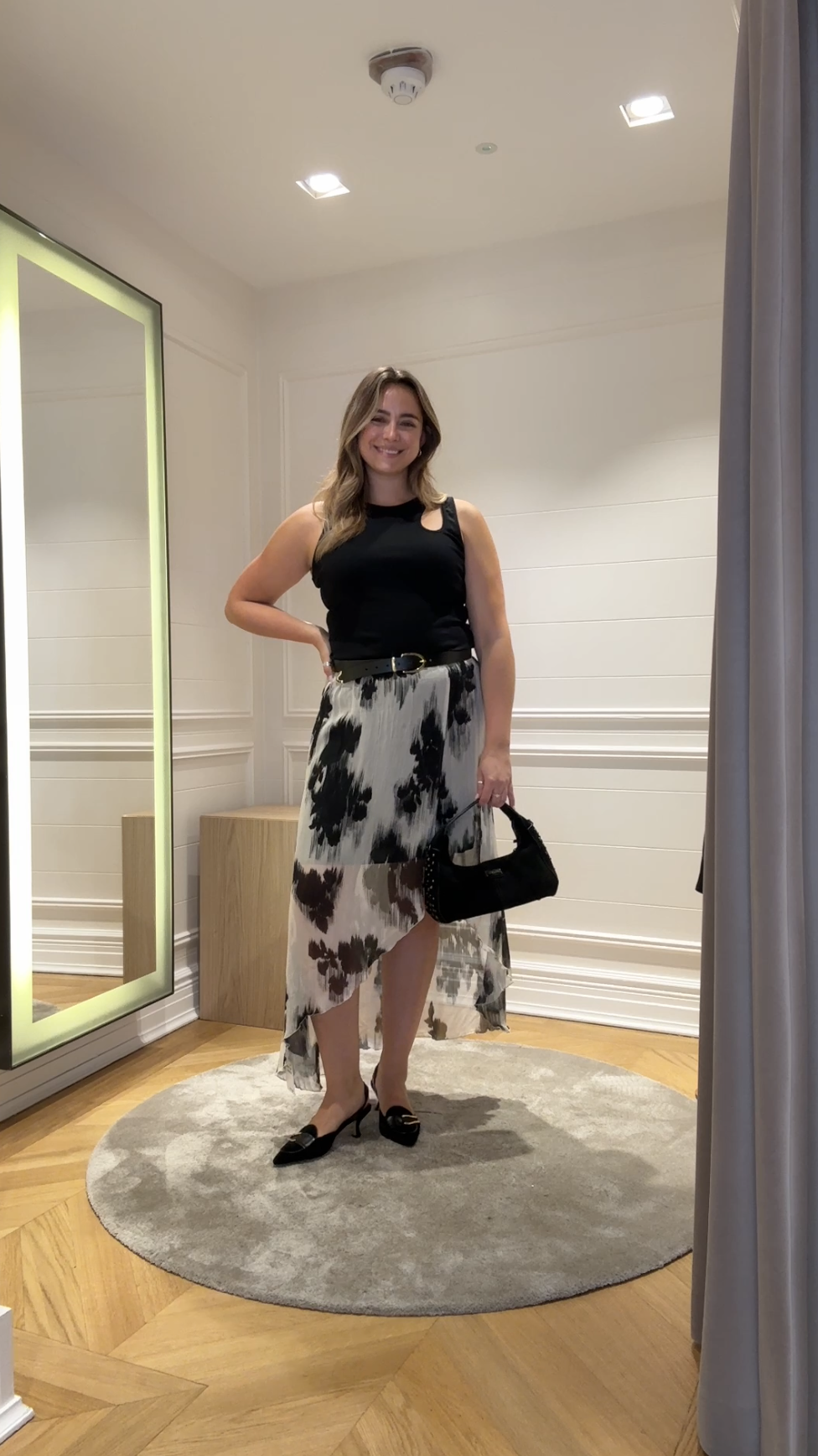 A size 14 model wearing the saint and sofia Lexi High Low midi skirt. A black and white floral design with a semi sheer finish and an asymmetric hem. It's paired with a black vest with a cut out detail at the shoulder.