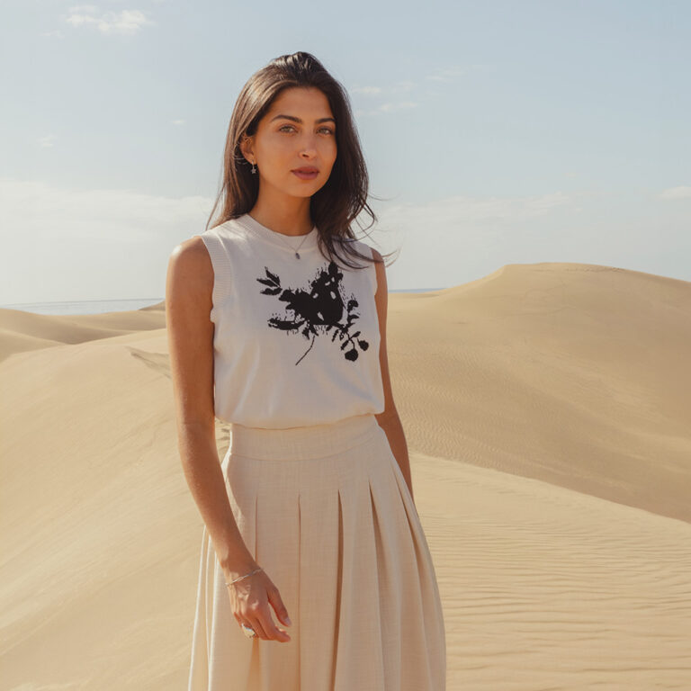 A model standing in the dunes wearing the Saint and Sofia Astra Sweater Vest with a linen midi skirt