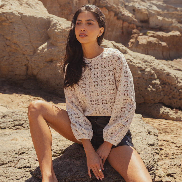 A model sat on a rocky terrain wearing a Saint and Sofia broderie anglaise long sleeve blouse with leather shorts.