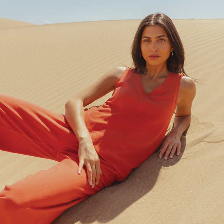 A model laying on the sand wearing a burnt orange linen co-ord by Saint and Sofia. A pair of wide leg trousers with a matching V-neck tunic top
