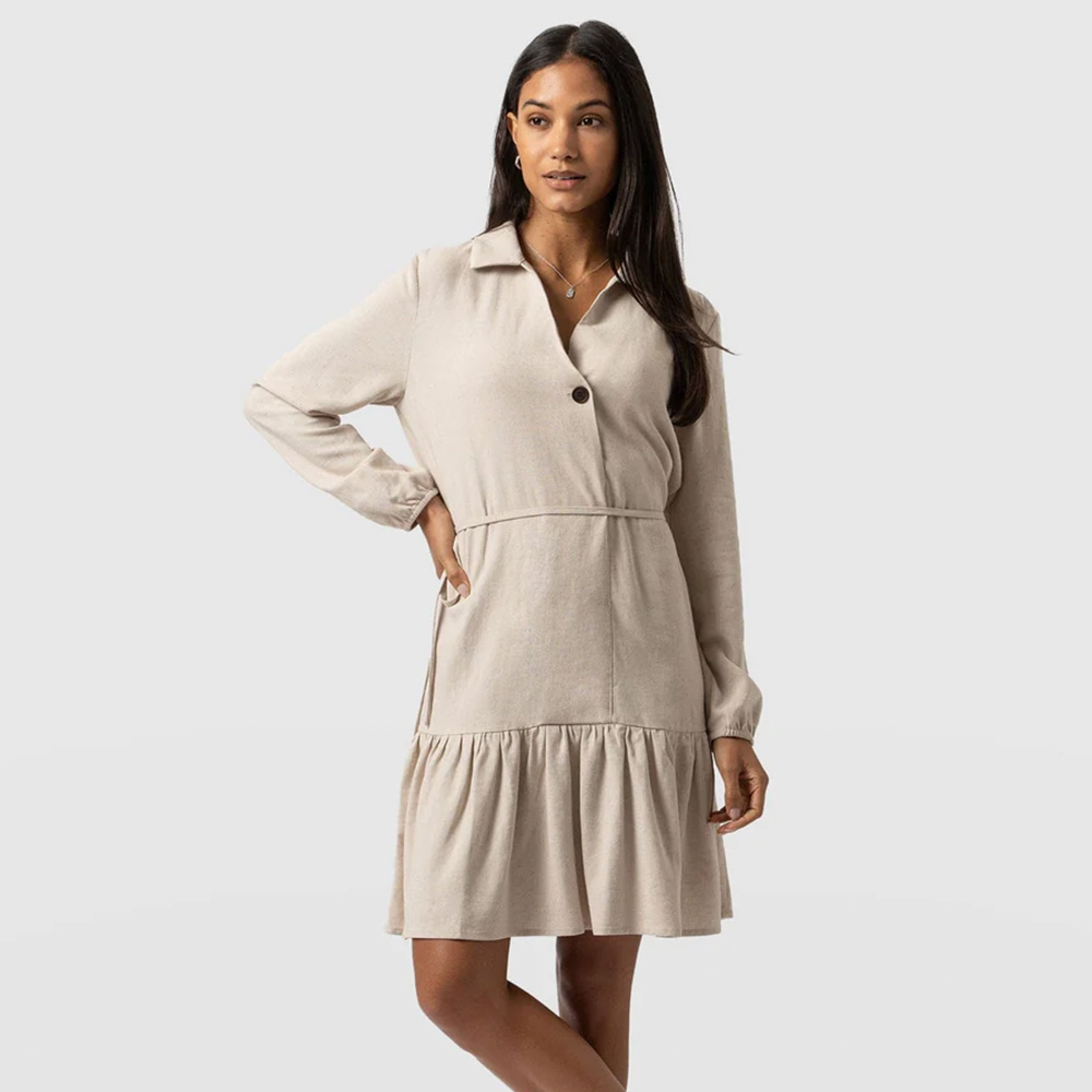 A model wearing the Saint and Sofia Aurelia Linen Mini Dress in oatmeal. A long sleeved design with a collared neckline, tired skirt, and self-tying belt at the waist.