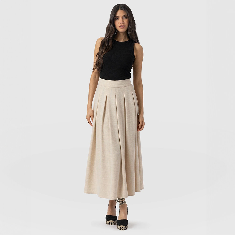 A model wearing the Saint and Sofia Eliza midi skirt. A pleated linen skirt in an oatmeal colour with a flat fronted waistband. Styled here with a black racer vest and black espadrille heels.