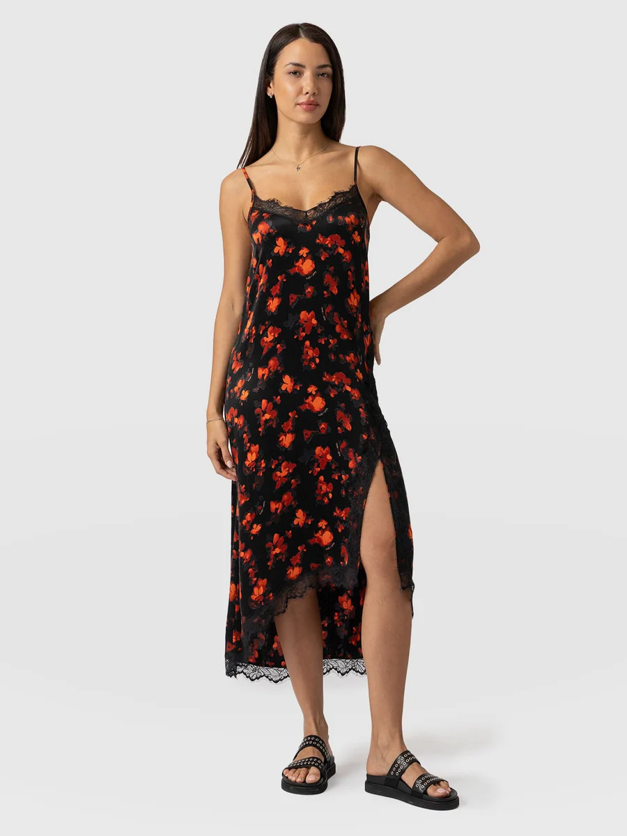 A model wearing the Saint and Sofia Harper Lace Cami Dress. A red and black wedding guest dress with lace trims and an asymmetric hemline.