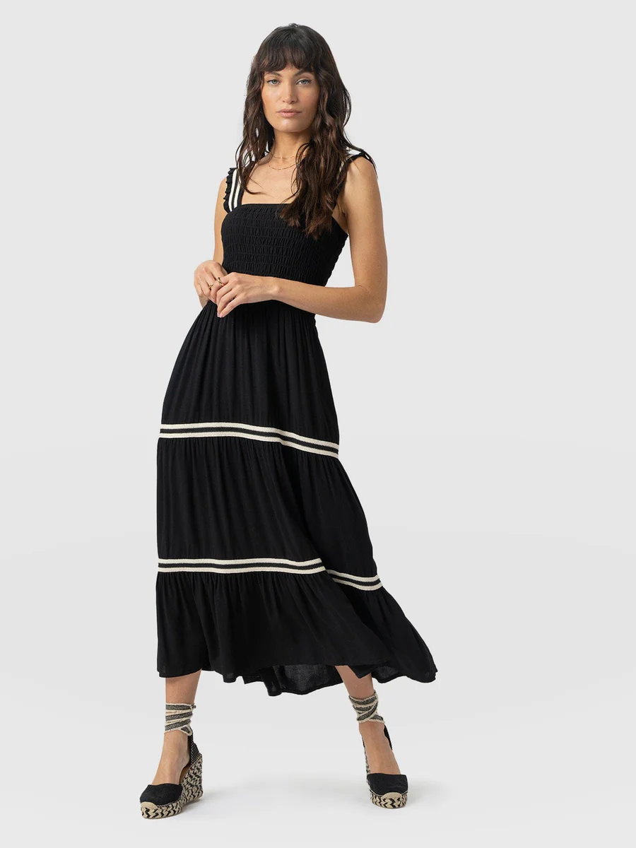 A model wearing the Saint + Sofia Maya shirring dress. A black sleeveless design with a ruched bodice and a tiered skirt split with contrasting white stripes. The perfect summer holiday dress.