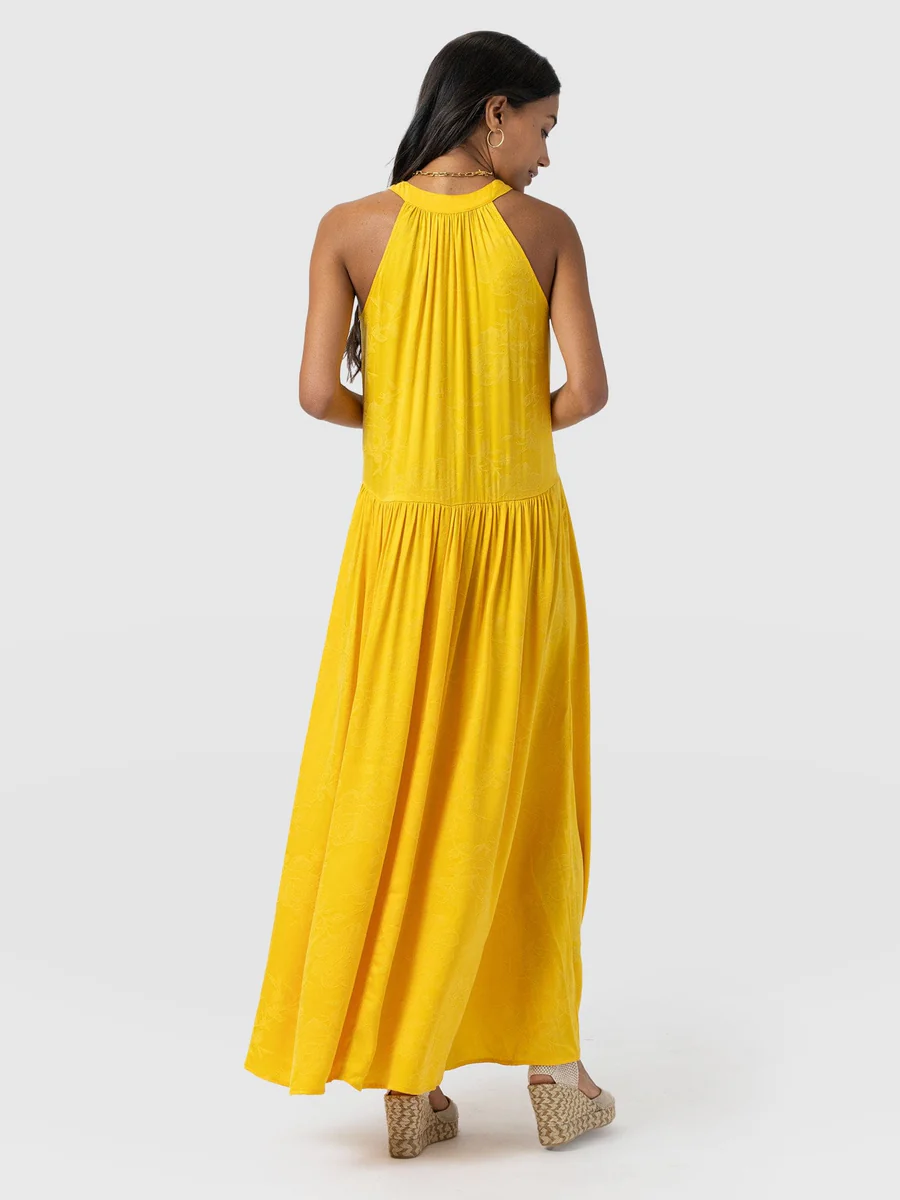 A model wearing the Saint and Sofia Zoey Halter Neck Dress. A beautifully wedding guest dress in a summery yellow with a floral jacquard texture and a cut out at the chest.