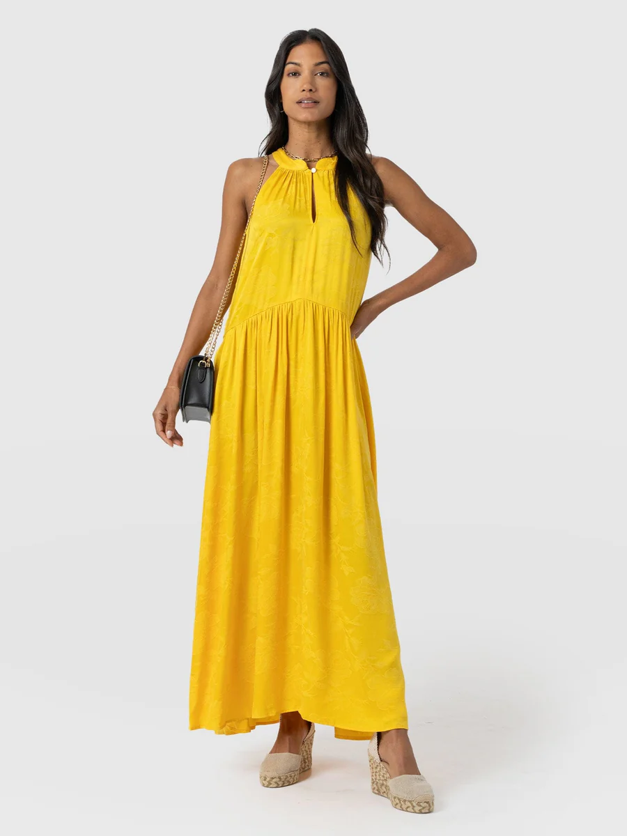 A model wearing the Saint and Sofia Zoey Halter Neck Dress. A beautifully wedding guest dress in a summery yellow with a floral jacquard texture and a cut out at the chest.