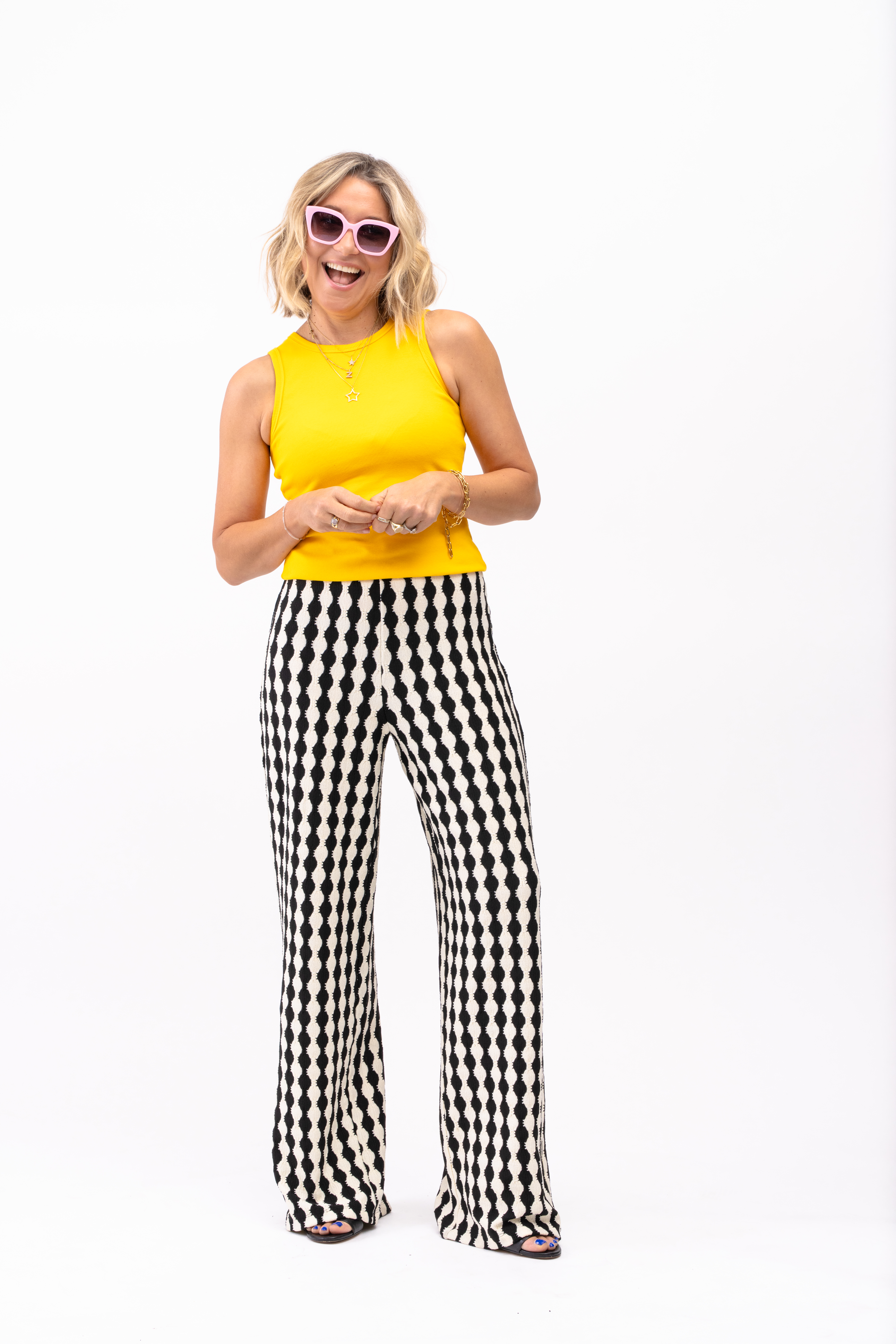 Zoë de Pass stood in a white studio wearing the Saint + Sofia Victory Wide Leg Trousers with the Nova Racer Vest in yellow. She wears pink sunglasses from her own brand.