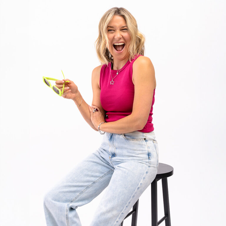 Zoe de Pass sat on a stool wearing a pink Saint and Sofia racer vest with the Saint and Sofia adjustable jeans and holding a pair of Zoe de Pass Eyewear in her hands