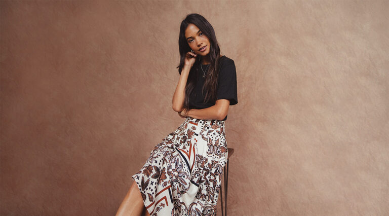 A model sat on a stool in front of a brown background wearing a black tee with the new Saint + Sofia Eliza pleated skirt in cream and brown paisley print.