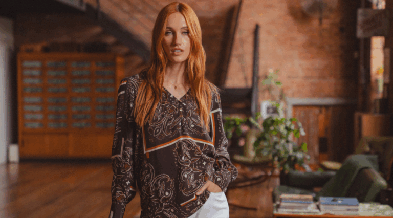 A model standing in a mid-century house wearing a brown paisley pattern long sleeve blouse with a V-neckline from Saint + Sofia.