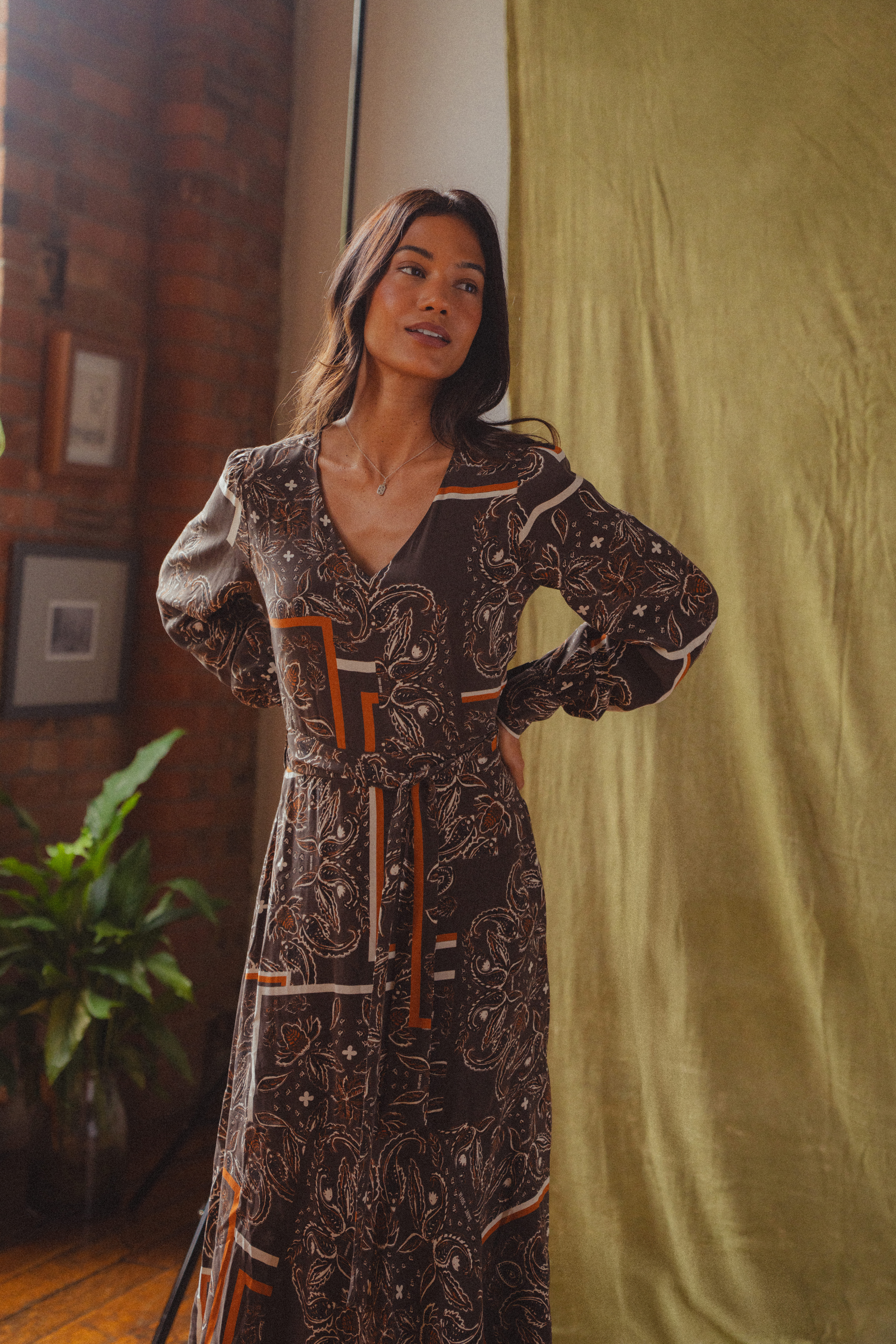 A model standing in a mid-century home wearing the Saint + Sofia Naya V Neck Dress in brown paisley.
