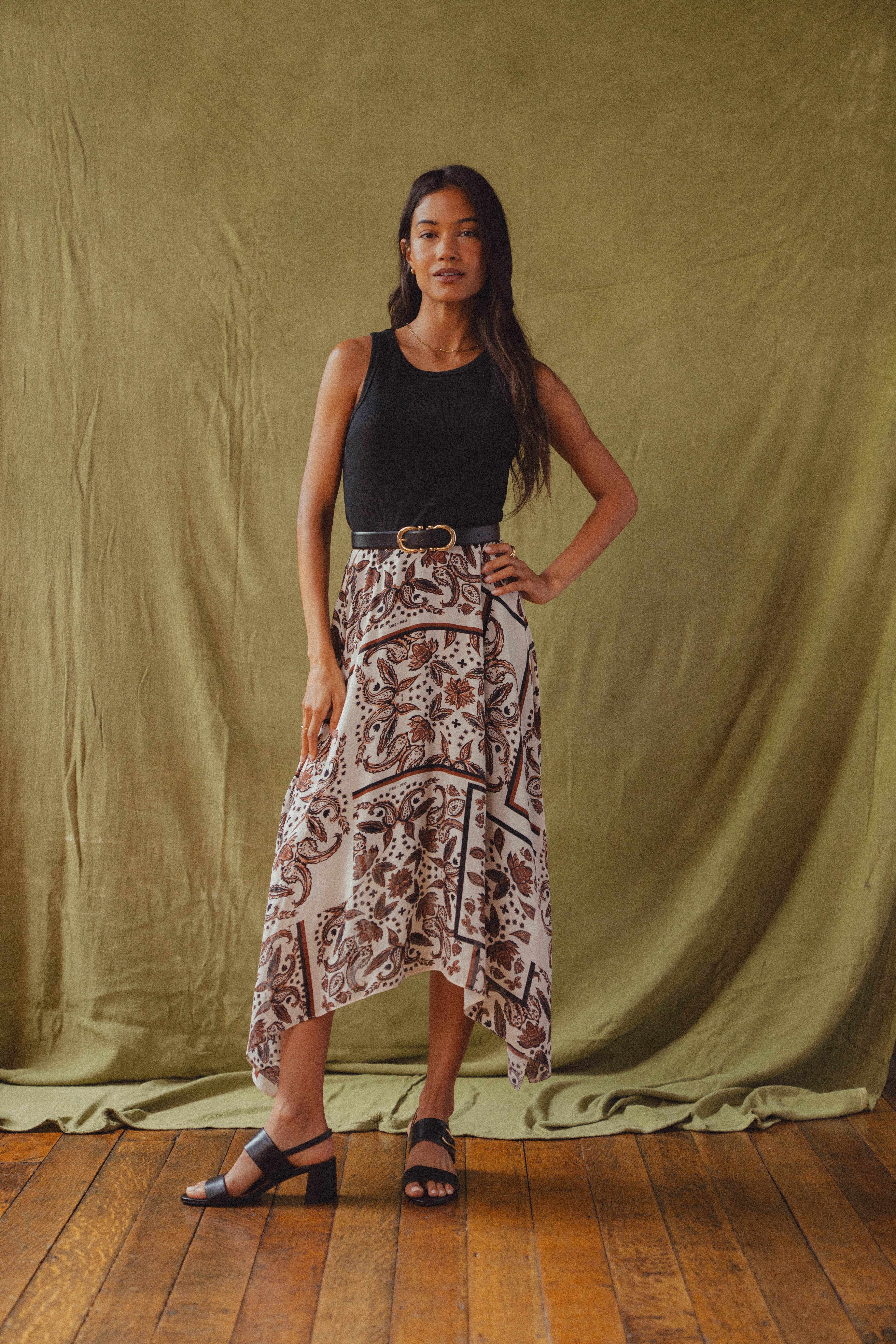 A model standing in front of a green back drop wearing the Saint + Sofia Lila Hanky Hem Dress with a black jersey bodice and a beige paisley pattern skirt.