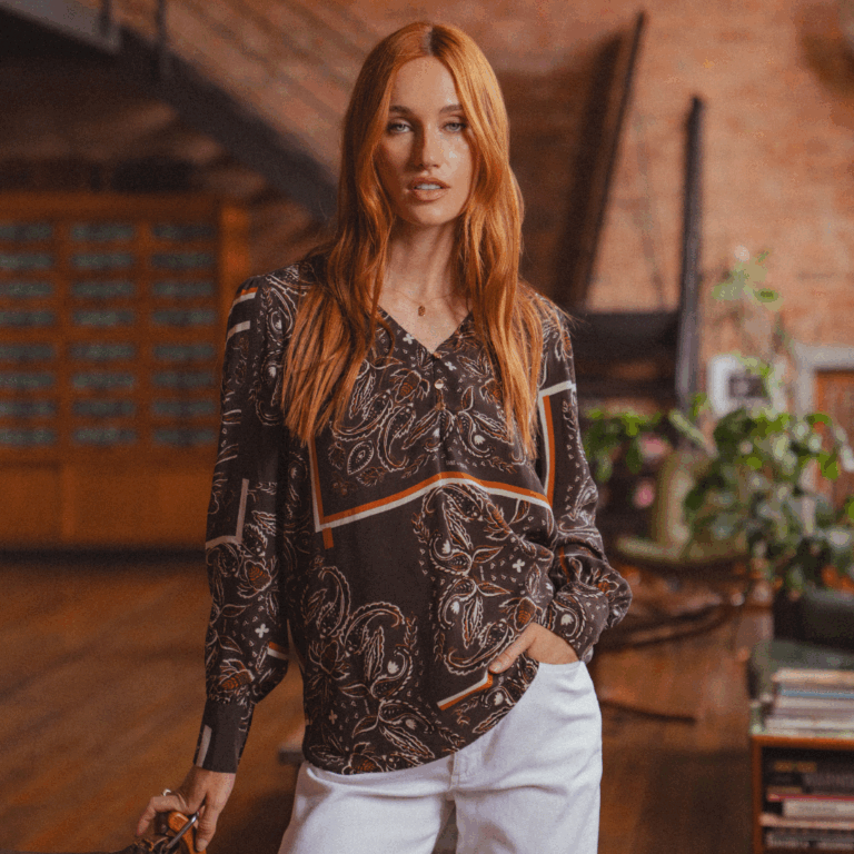 A model standing in a mid-century house wearing a brown paisley pattern long sleeve blouse with a V-neckline from Saint + Sofia.
