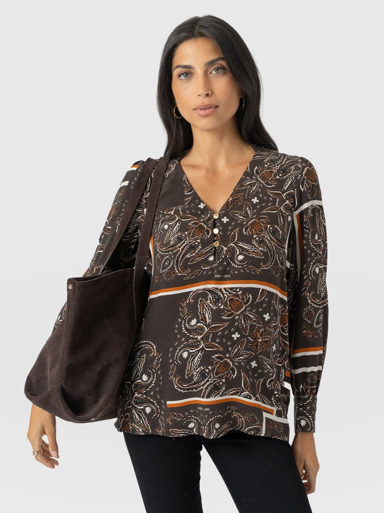 A model standing in front of a white backdrop wearing the Saint + Sofia Joselyn Blouse in a brown paisley pattern