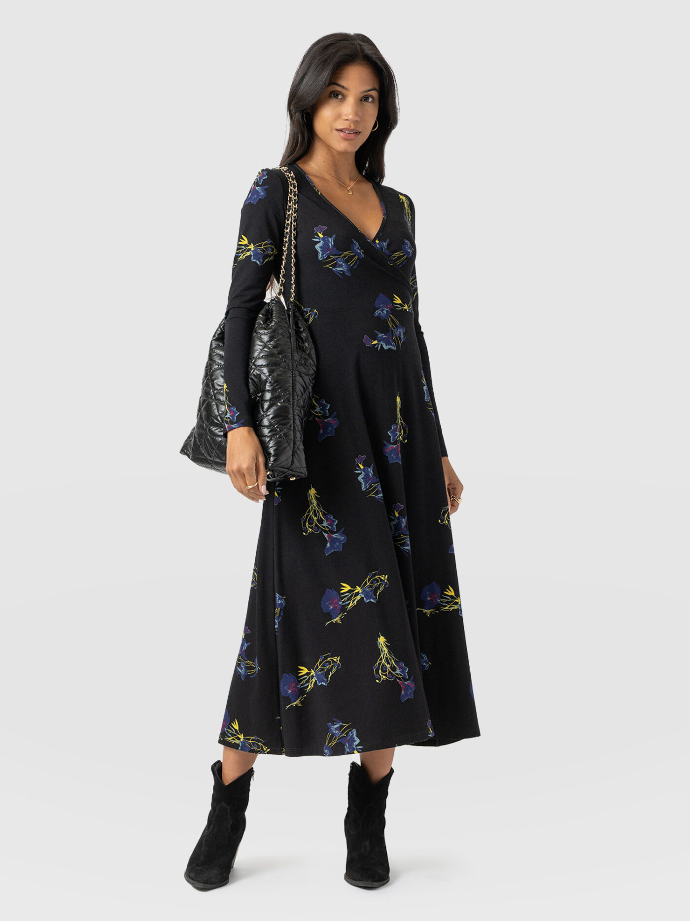 A model wearing the Saint and Sofia Maggie Wrap Dress. A long-sleeved design with a fixed wrap bodice that's perfect for flattering apple body shapes. Here, it is styled with a quilted nylon shoulder bag and black suede ankle boots.