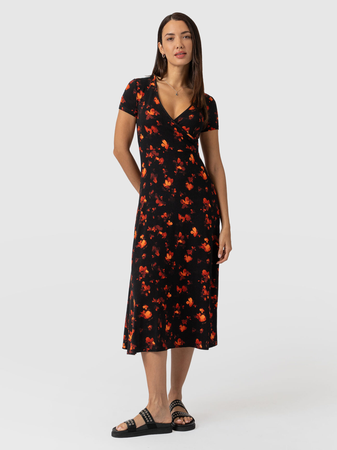 A model standing the Saint and Sofia Maggie Wrap Dress. She styles it with a pair of sliders. This mock wrap dress has short sleeves and features a mock wrap bodice with a V neckline.
