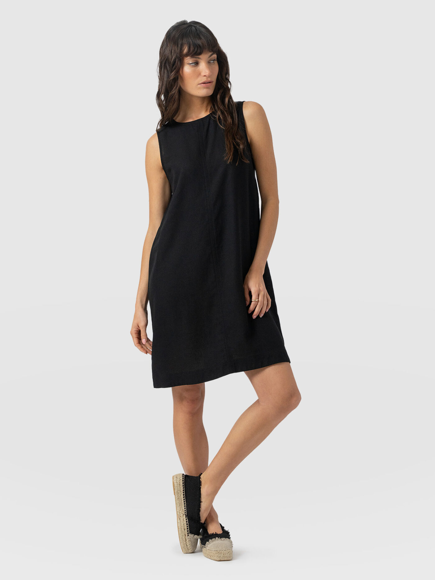 A model wearing the Saint and Sofia Corinne Shift Dress in black. A crew neck, sleeveless mini that falls loosely over your figure and is perfect for appl body shapes.