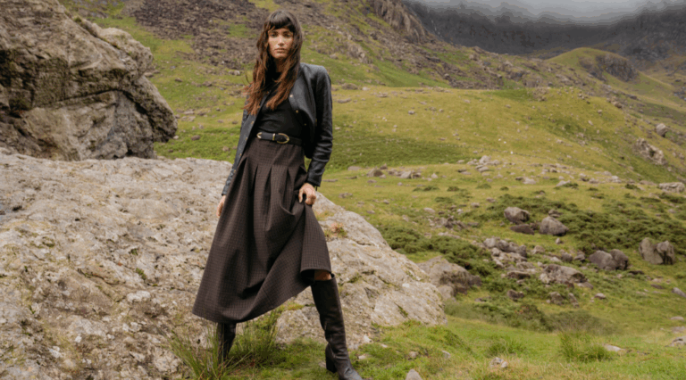 A model standing against a grassy hill backdrop beside a grey rock wearing the Saint and Sofia Macie Zip Up Dress and a leather jacket