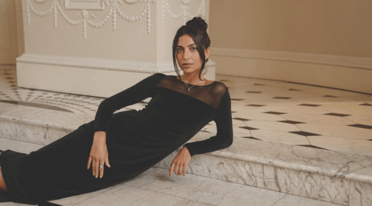 A model lounging on marbled steps wearing a black, long-sleeved body con dress with a semi-sheer mesh panel at the decolletage.