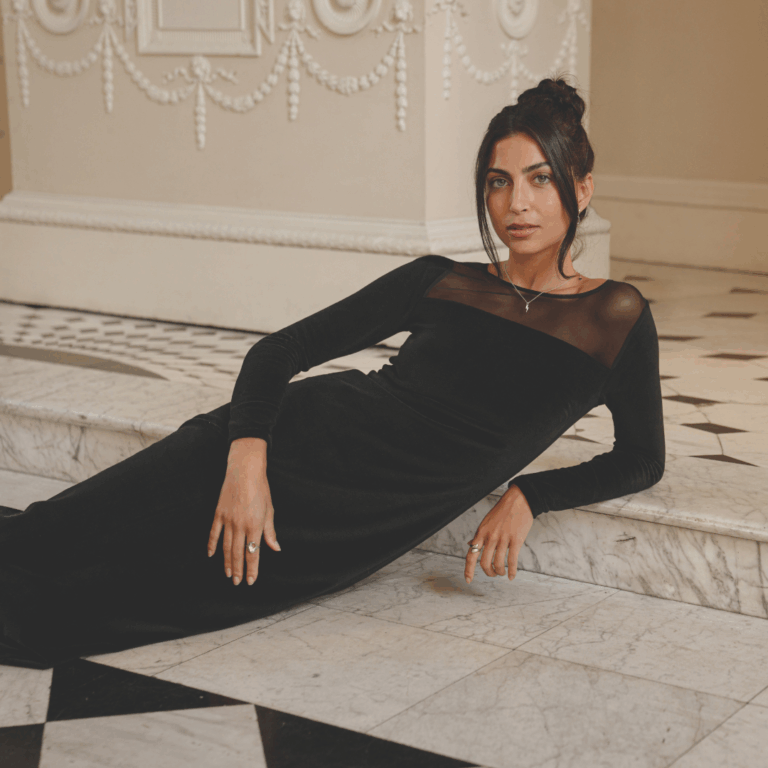 A model lounging on marbled steps wearing a black, long-sleeved body con dress with a semi-sheer mesh panel at the decolletage.