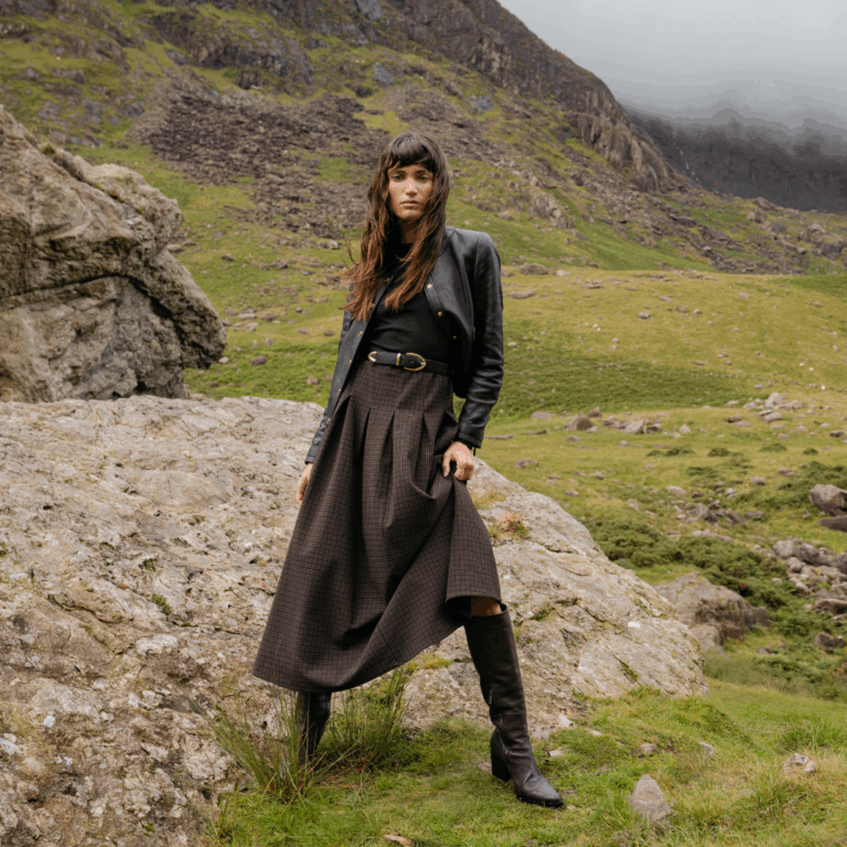 A model standing against a grassy hill backdrop beside a grey rock wearing the Saint and Sofia Macie Zip Up Dress and a leather jacket