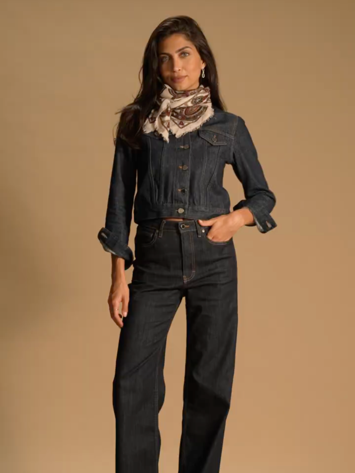 A model standing in front of a brown background wearing a denim jacket, matching wide leg jeans and a beige paisley neck tie by Saint and Sofia.