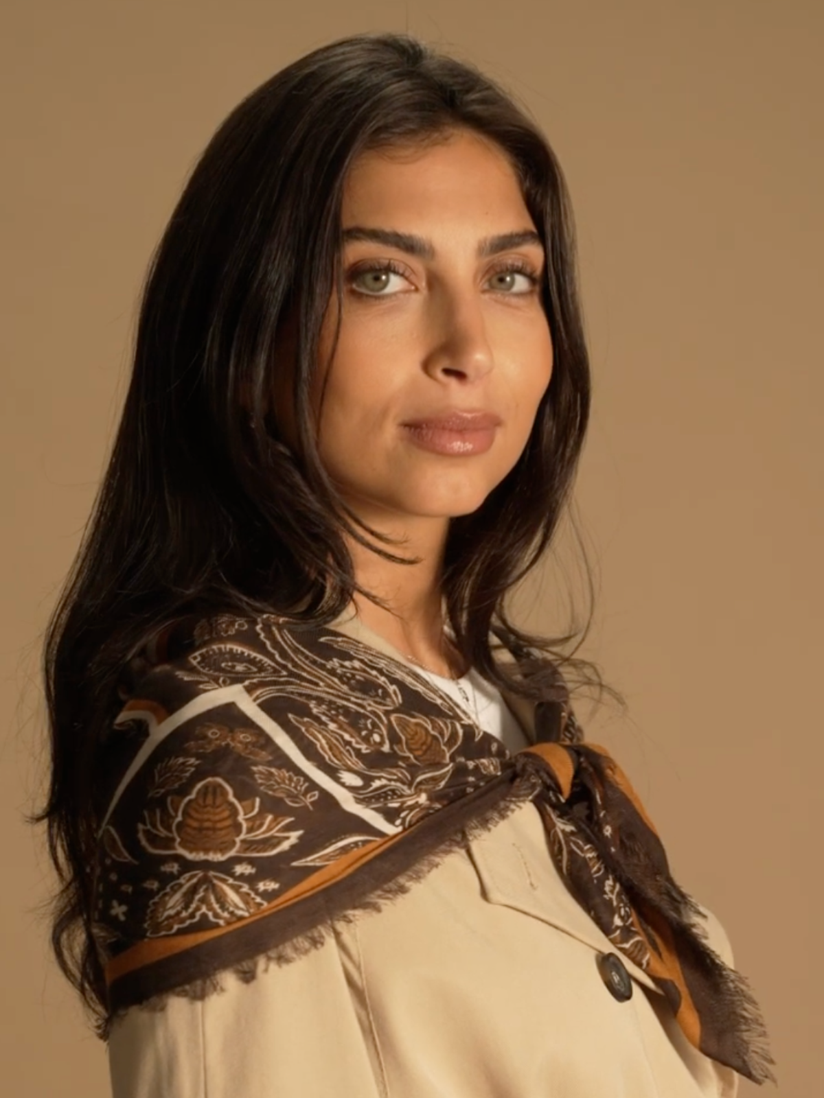 A close up shot of a model standing to the side, twisting her head to face the camera. She wears a camel trench coat and over her shoulders she wears a brown paisley scarf knotted at the chest. 