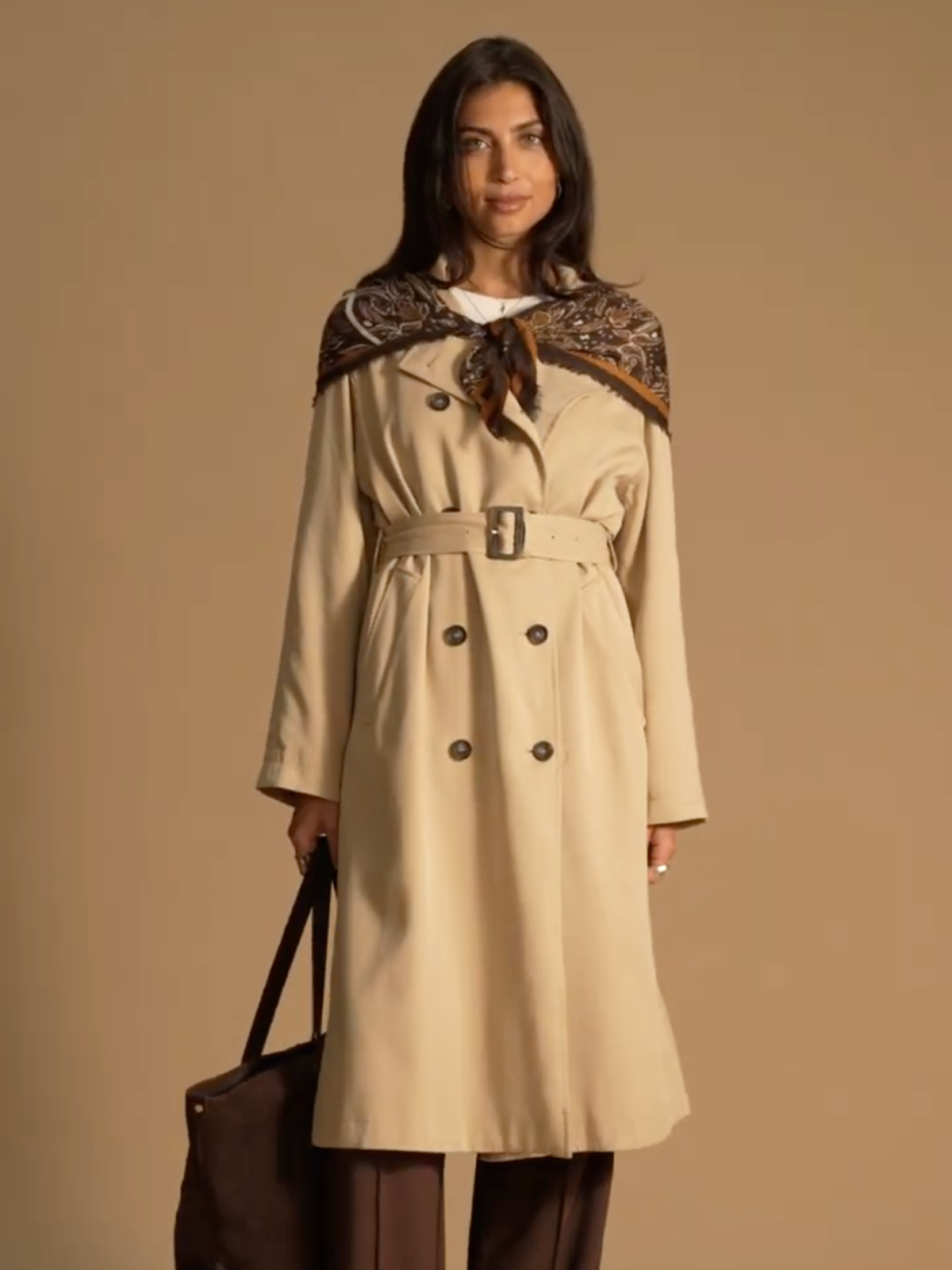 A model standing in front of a brown background wearing a camel trench coat with a brown tote bag in her head. She has a brown paisley scarf wrapped around her shoulders and fastened at her chest as a shawl.